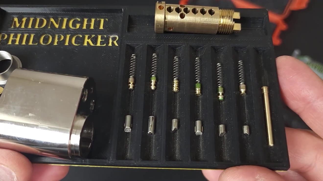A euro cylinder taken completely apart and laid out in order on a tray: the brass plug with six chambers, six springs, six spool driver pins, six key pins, and the shell, held in Isaac&rsquo;s hands